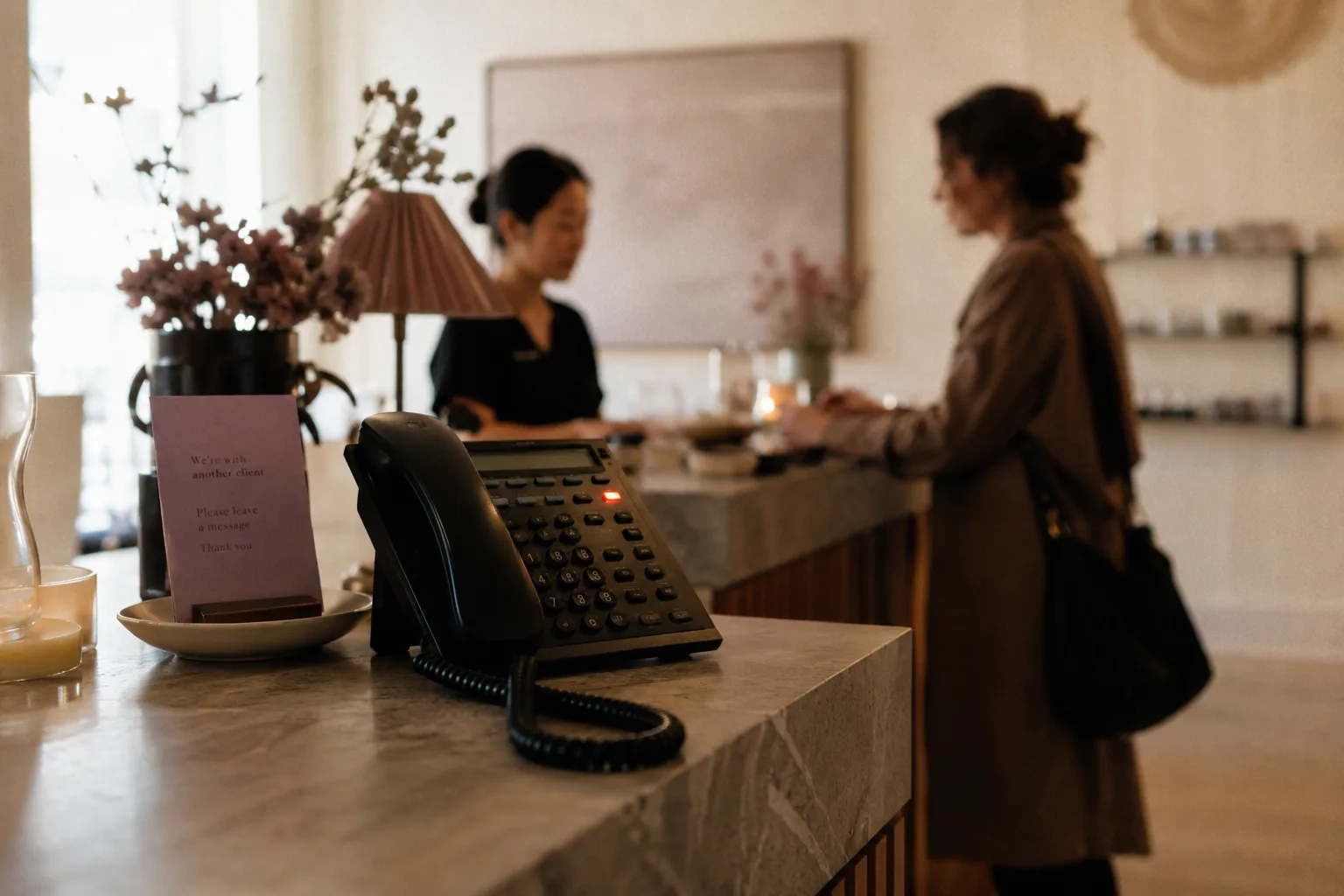A front-desk phone rings unattended while the receptionist helps another client; a small sign reads 'We're with another client — please leave a message.'